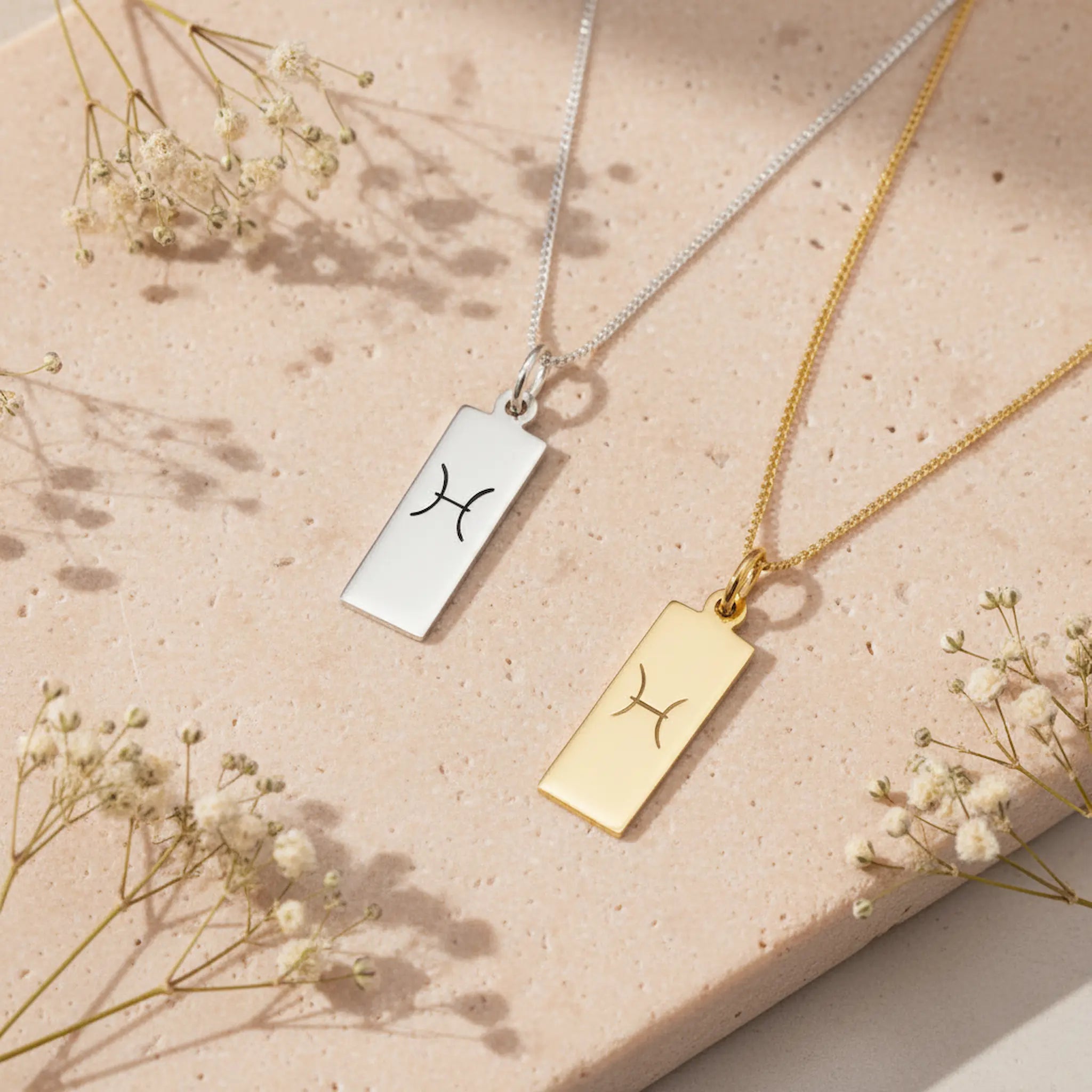 two pisces necklaces in silver and gold featuring rectangle pendants engraved with a minimalist pisces zodiac star sign line glyph symbol against a light stone surface