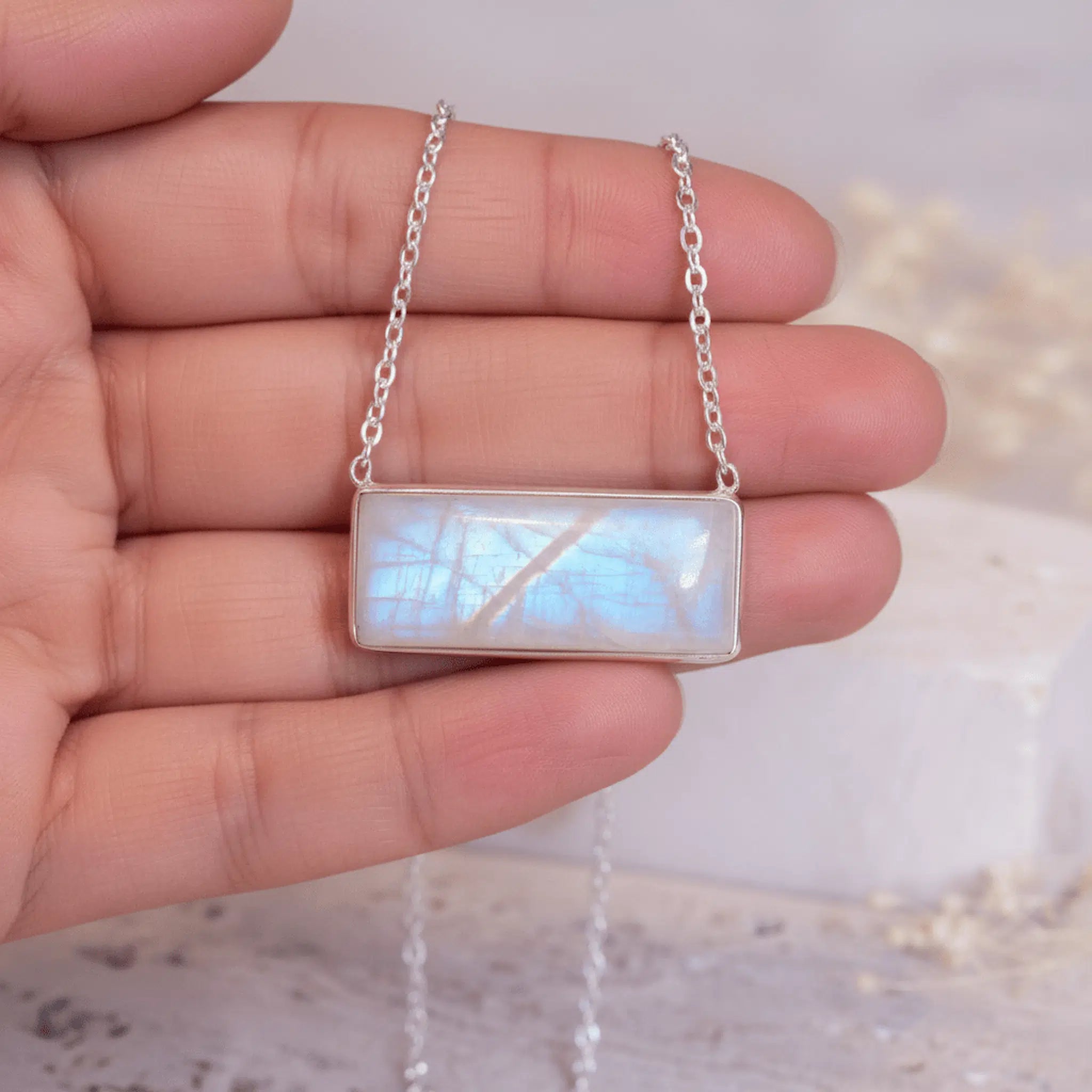 Hand holding a silver necklace with a rectangular moonstone pendant against a blurred background