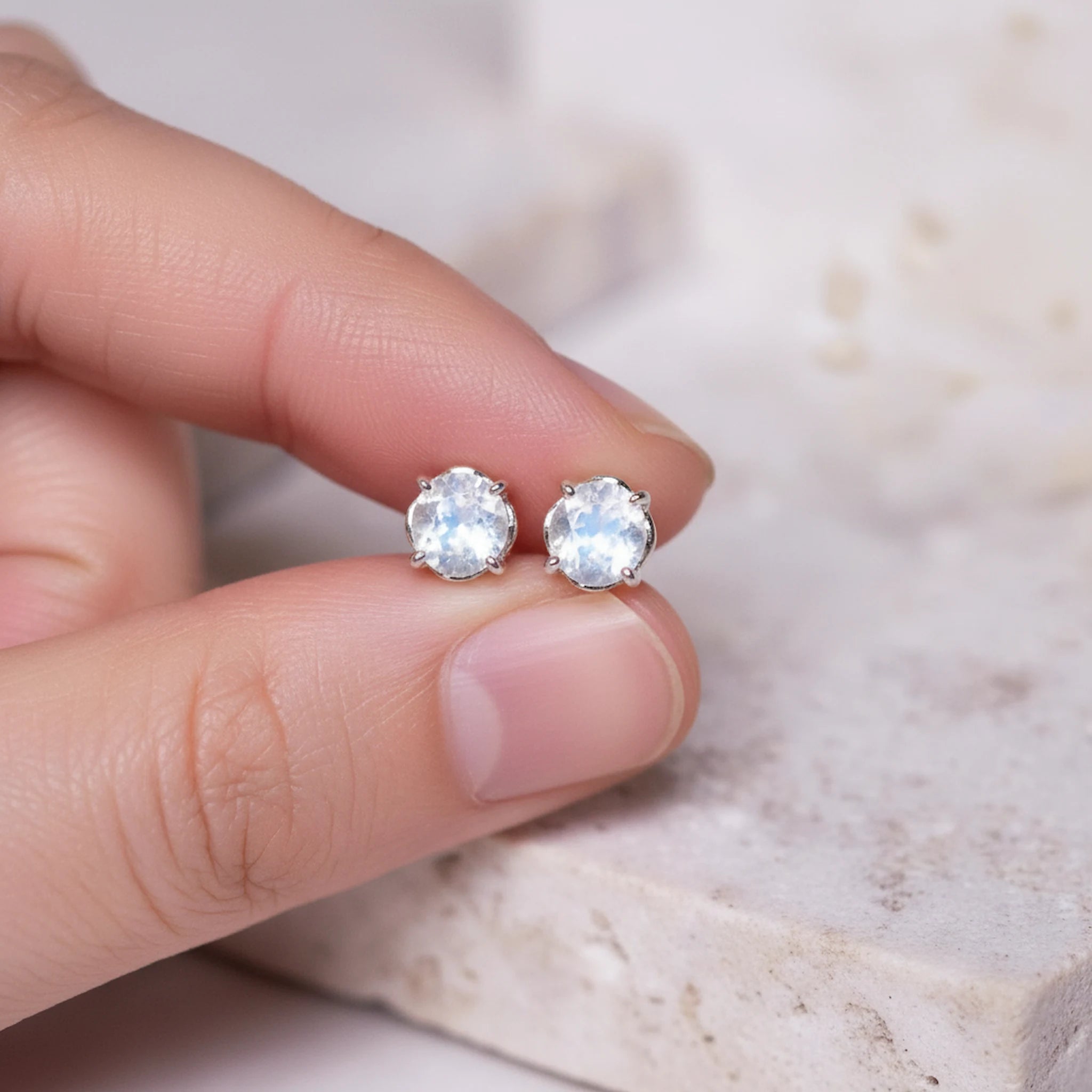 Silver stud earrings with natural rainbow moonstone gemstones held between fingers on a neutral background