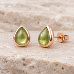 pair of august birthstone natural peridot earrings - Teardrop-shaped stud earrings with smooth olive green gemstones in polished rose gold settings, displayed on a textured beige stone surface.