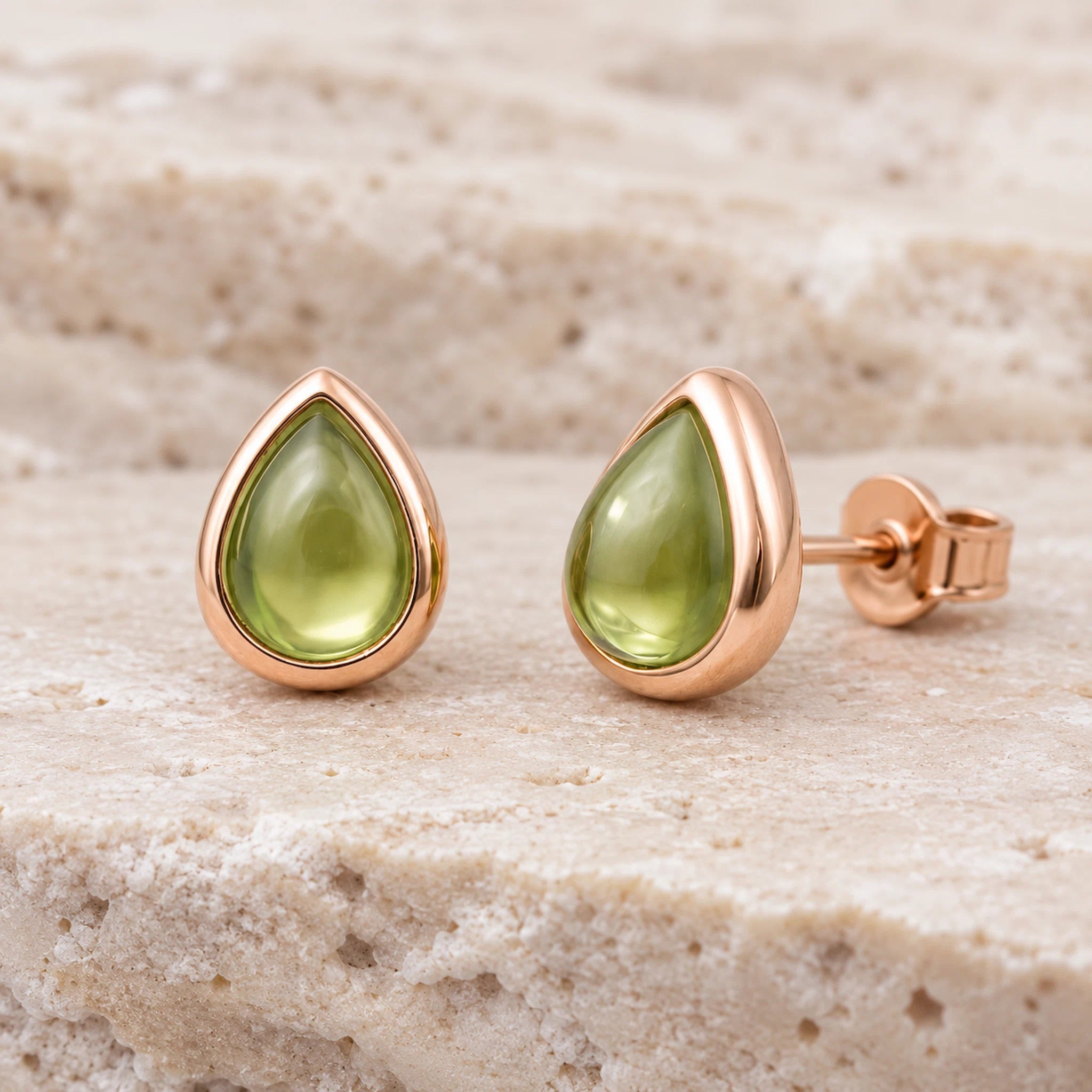 pair of august birthstone natural peridot earrings - Teardrop-shaped stud earrings with smooth olive green gemstones in polished rose gold settings, displayed on a textured beige stone surface.