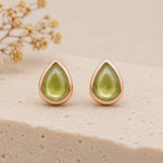 A pair of august birthstone natural peridot earrings - teardrop-shaped stud earrings with smooth light green stones in a polished rose gold setting, displayed on a textured beige stone surface with dried flowers in the background.
