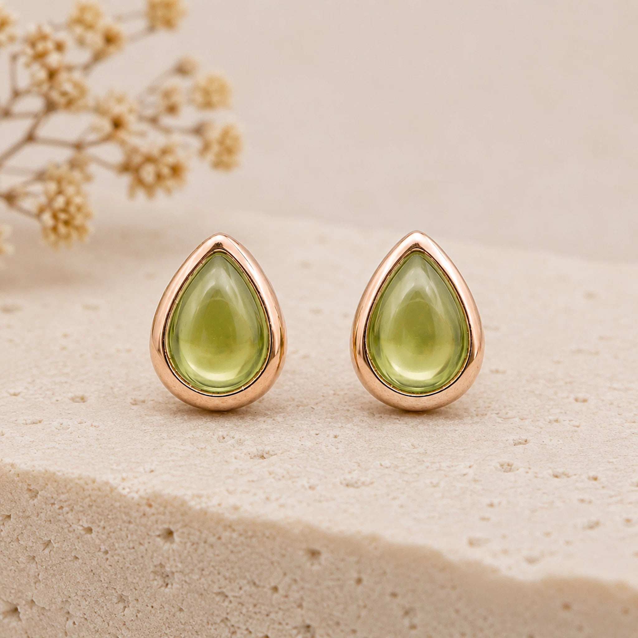 A pair of august birthstone natural peridot earrings - teardrop-shaped stud earrings with smooth light green stones in a polished rose gold setting, displayed on a textured beige stone surface with dried flowers in the background.