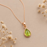 A rose gold august birthstone necklace with a teardrop-shaped green natural peridot gemstone pendant, displayed on a neutral beige background with dried white flowers.