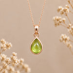 A rose gold August Birthstone Necklace with a teardrop pendant with a green peridot gemstone, suspended from a delicate rose gold chain against a warm beige background with blurred dried flowers.
