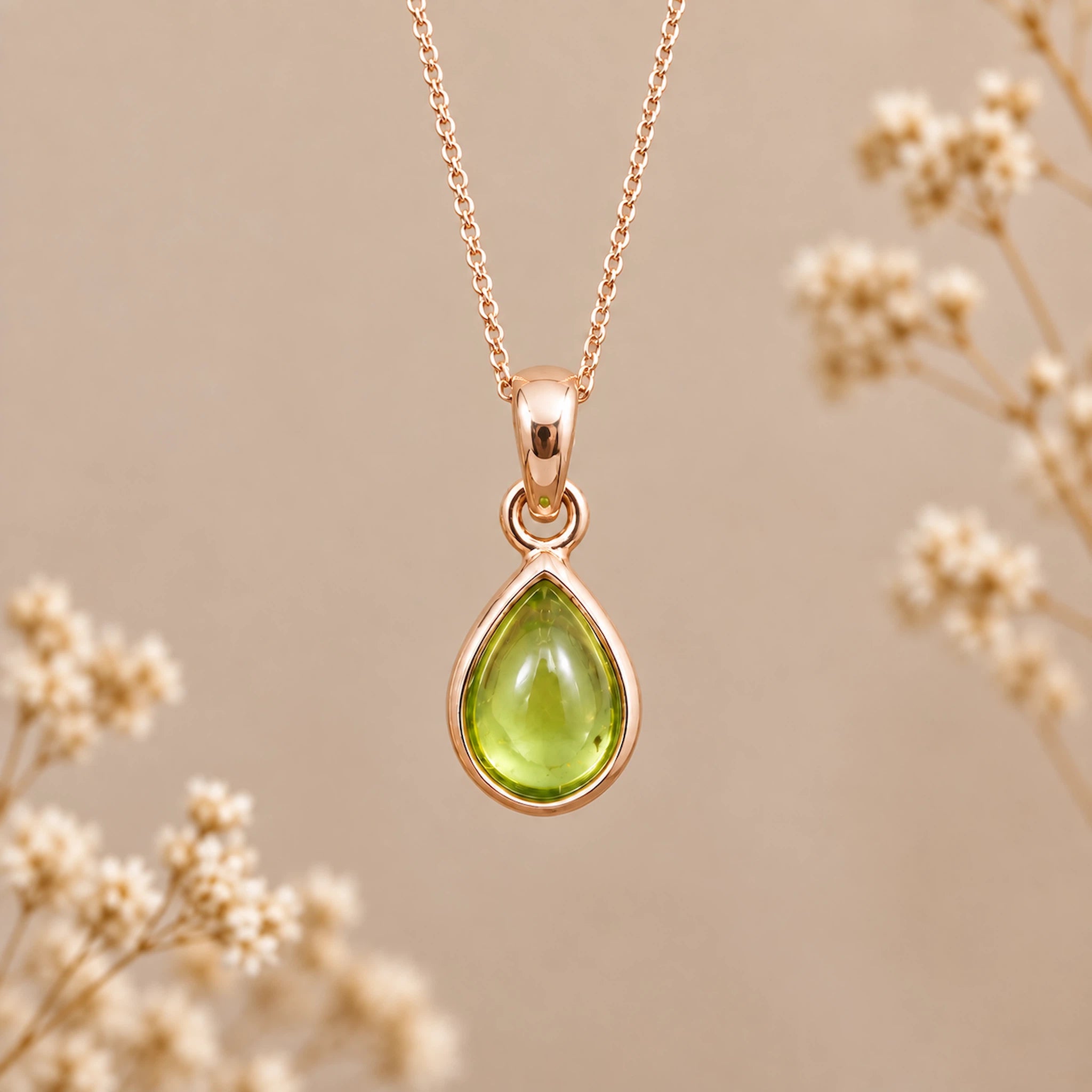 A rose gold August Birthstone Necklace with a teardrop pendant with a green peridot gemstone, suspended from a delicate rose gold chain against a warm beige background with blurred dried flowers.