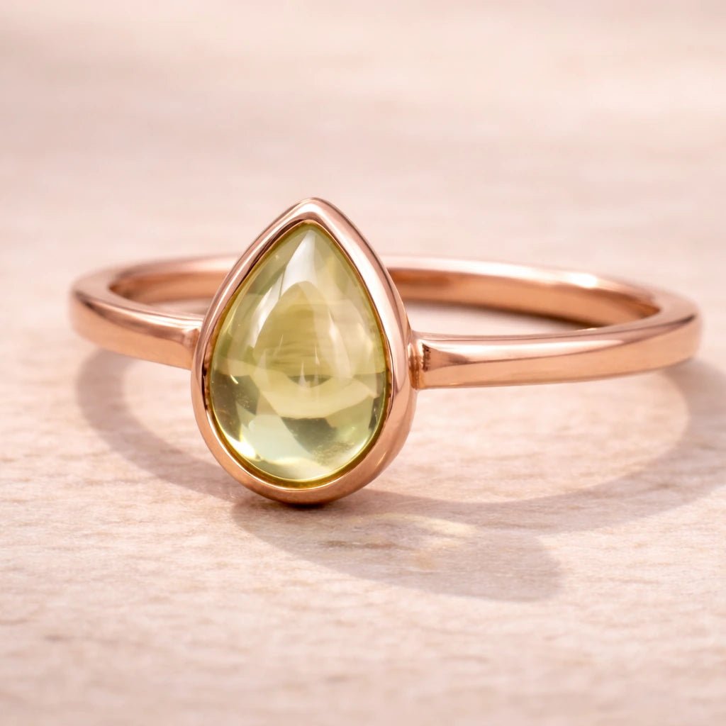 Rose gold plated August birthstone ring with a pear-shaped light green natural peridot gemstone cabochon in a polished low-profile bezel setting on a beige stone surface