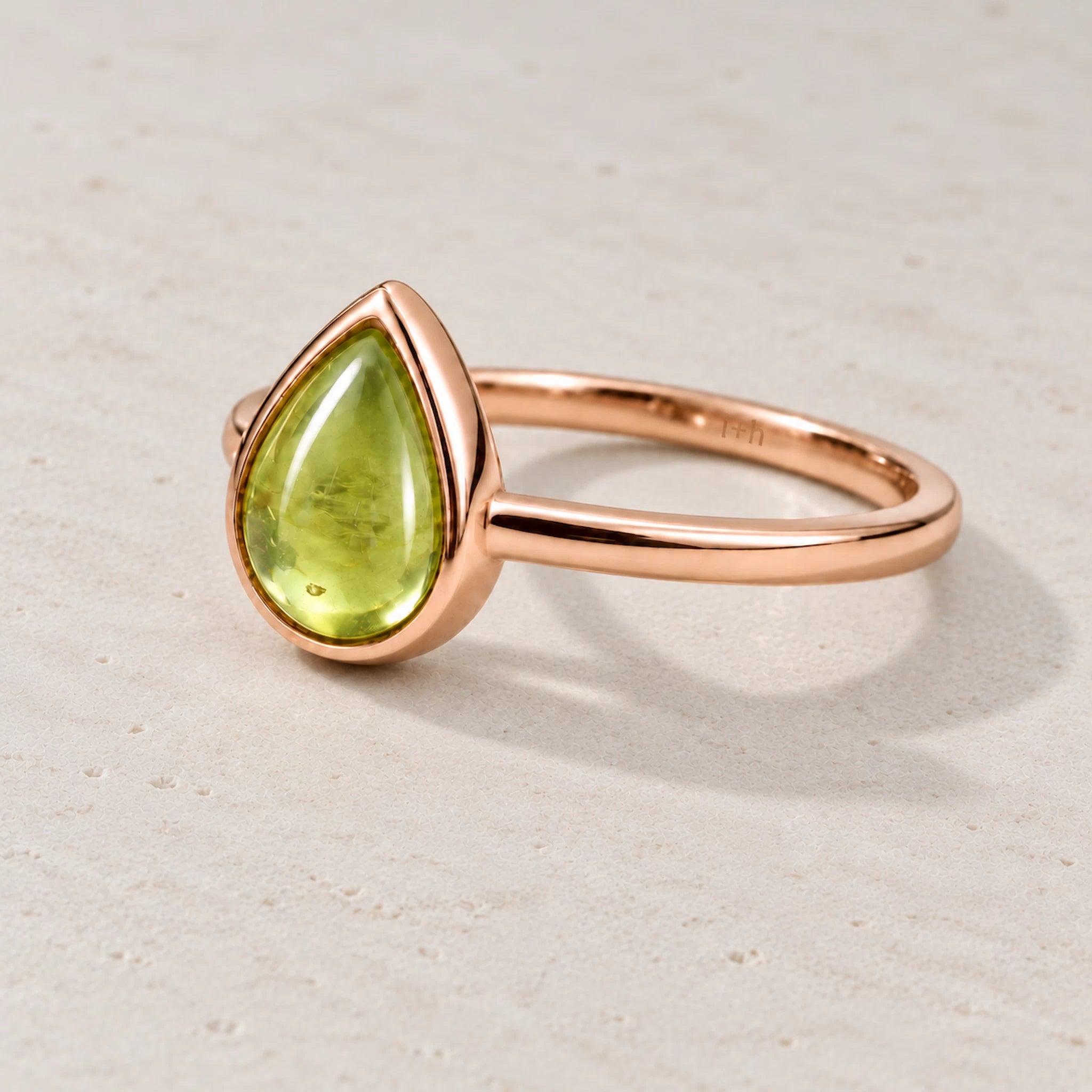 Rose gold plated August birthstone ring with a pear-shaped light green natural peridot gemstone cabochon in a polished low-profile bezel setting on a beige stone surface