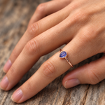 A close-up of a hand wearing a rose gold ring with a natural tanzanite gemstone, resting on a rustic wooden surface.