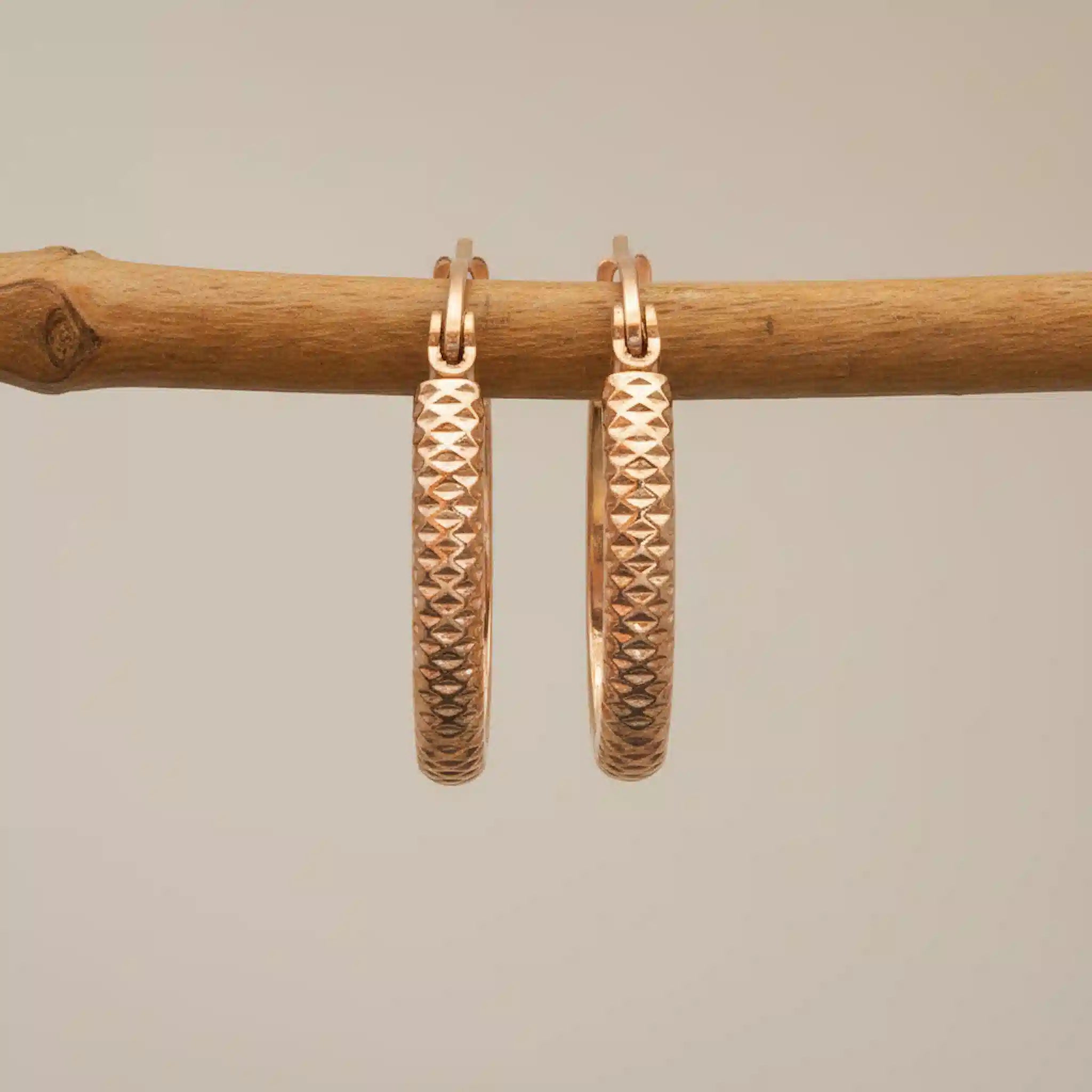 Rose gold hoop earrings on a wooden stick against a beige background