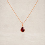 A rose gold chain necklace with a january birthstone teardrop-shaped dark red natural garnet gemstone pendant, displayed against a neutral textured beige background.