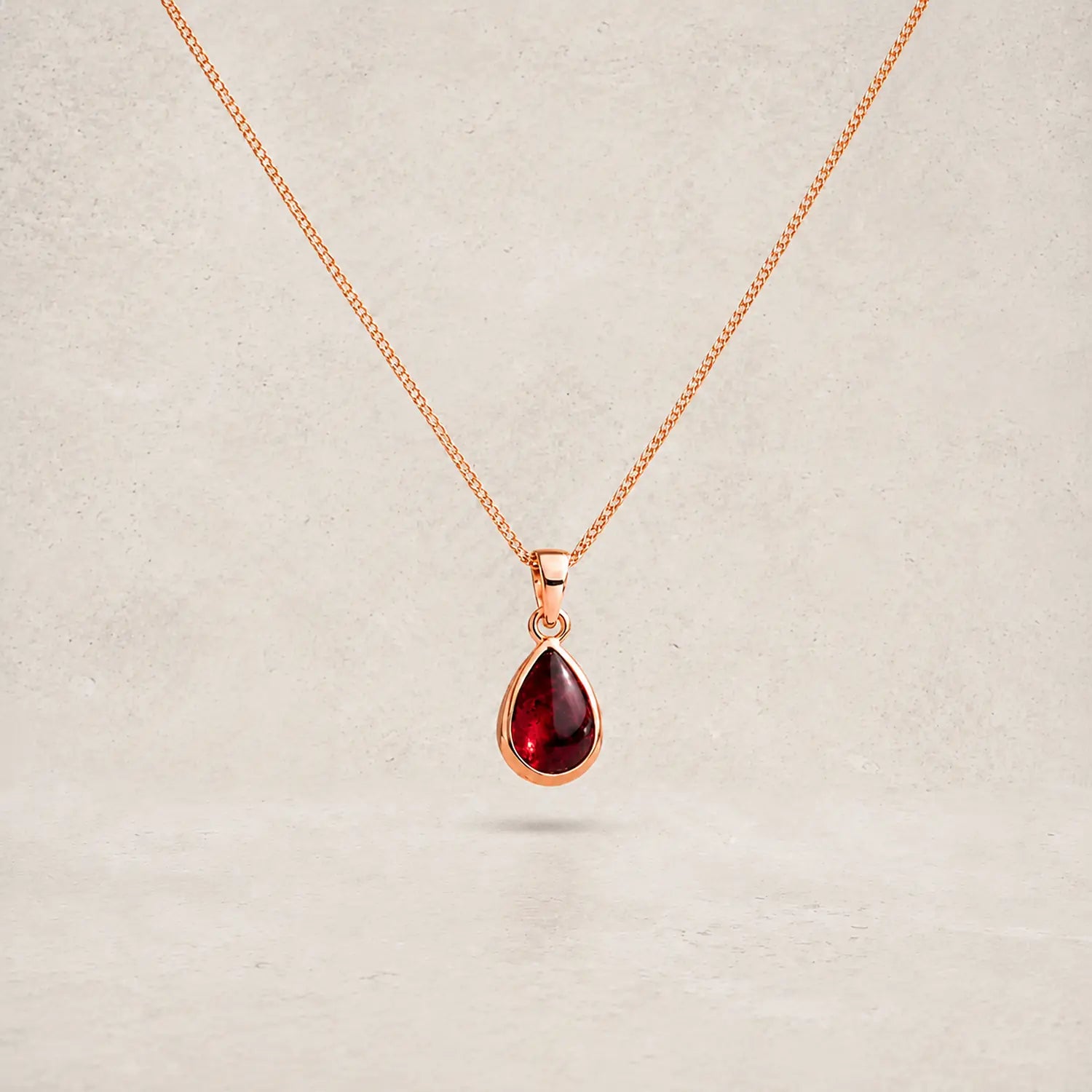 A rose gold chain necklace with a january birthstone teardrop-shaped dark red natural garnet gemstone pendant, displayed against a neutral textured beige background.