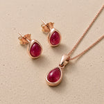 Rose gold July Birthstone Jewellery set featuring a teardrop pendant necklace and matching stud earrings with oval-cut deep red natural ruby gemstones, displayed on a textured beige surface.