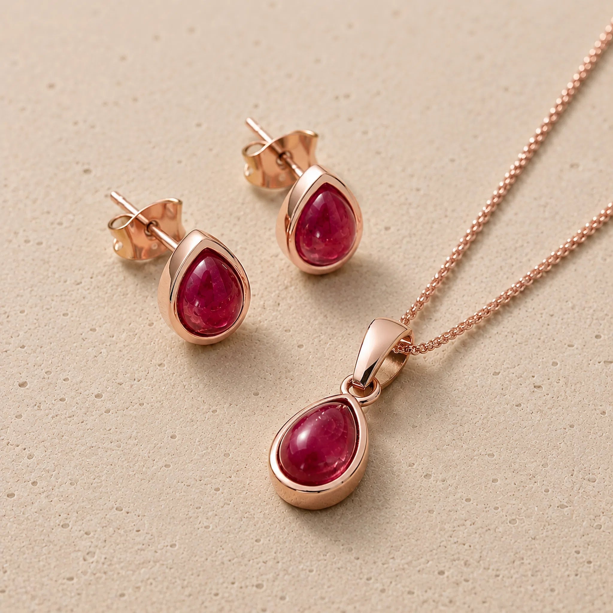 Rose gold July Birthstone Jewellery set featuring a teardrop pendant necklace and matching stud earrings with oval-cut deep red natural ruby gemstones, displayed on a textured beige surface.