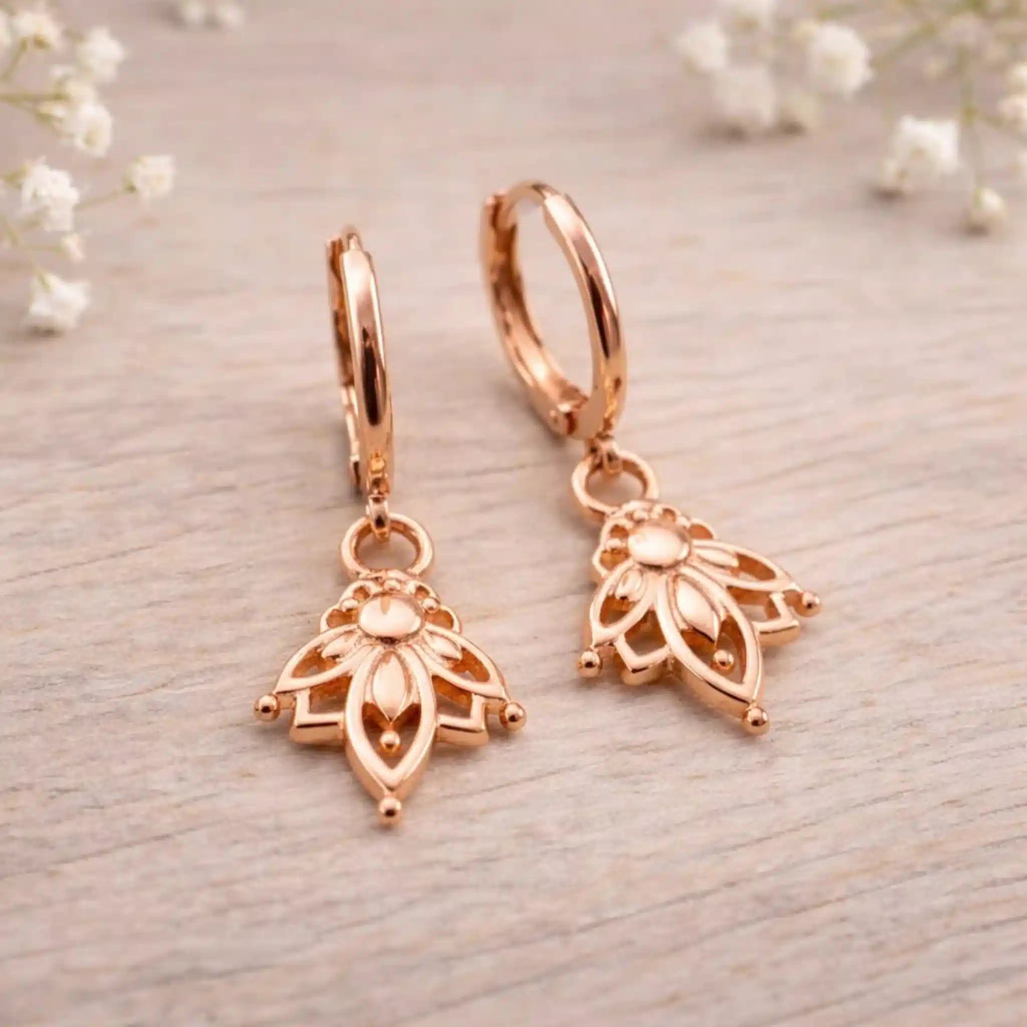 Rose gold hoop earrings with lotus flower floral designs on a light wooden surface.