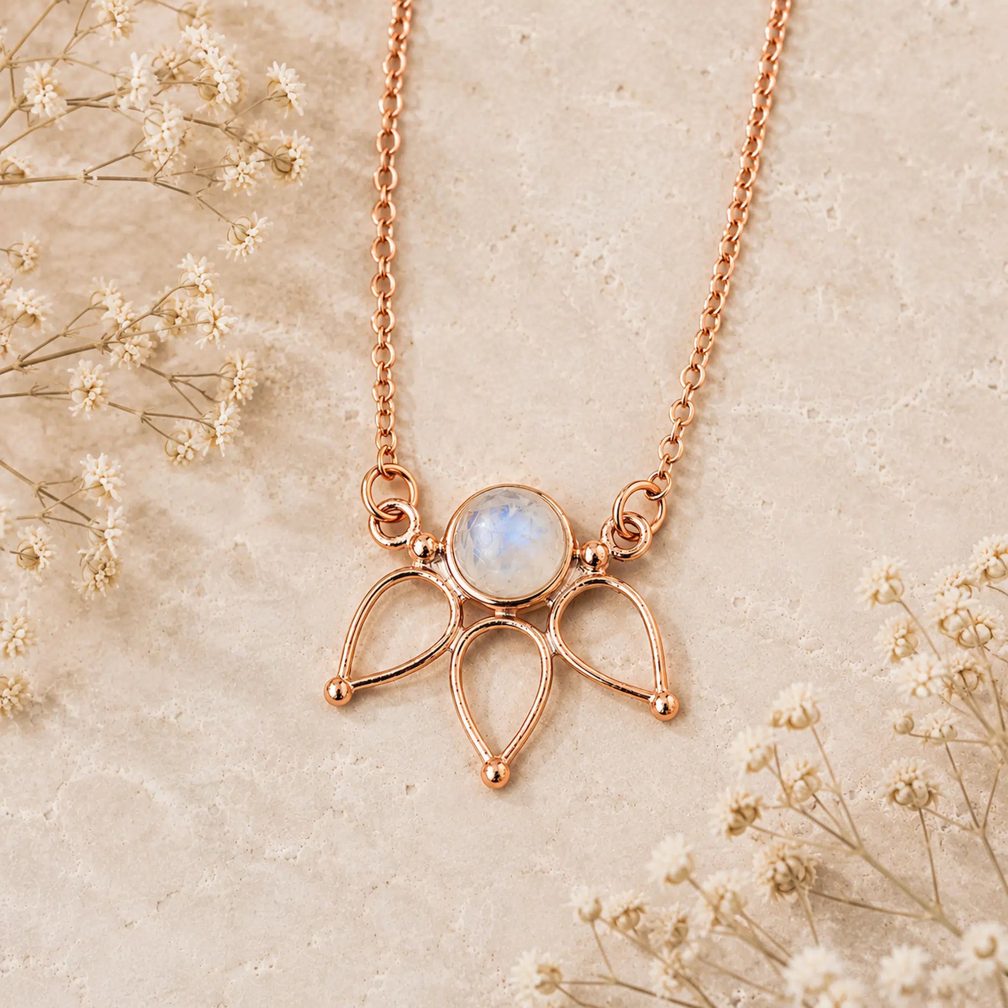 A rose gold necklace featuring a delicate three-pronged pendant with a round iridescent moonstone in the center, displayed on a textured beige surface with dried baby's breath flowers.