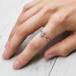Hand wearing a rose gold march birthstone ring with a blue aquamarine gemstone on a light wooden surface