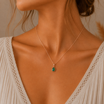 A close-up of a woman wearing a delicate rose gold chain may birthstone necklace with a small teardrop-shaped natural emerald green pendant, styled with a cream-colored lace top.