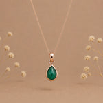 A rose gold may birthstone necklace with a teardrop pendant necklace with a natural green emerald gemstone, displayed against a warm beige background with small blurred dried flowers.