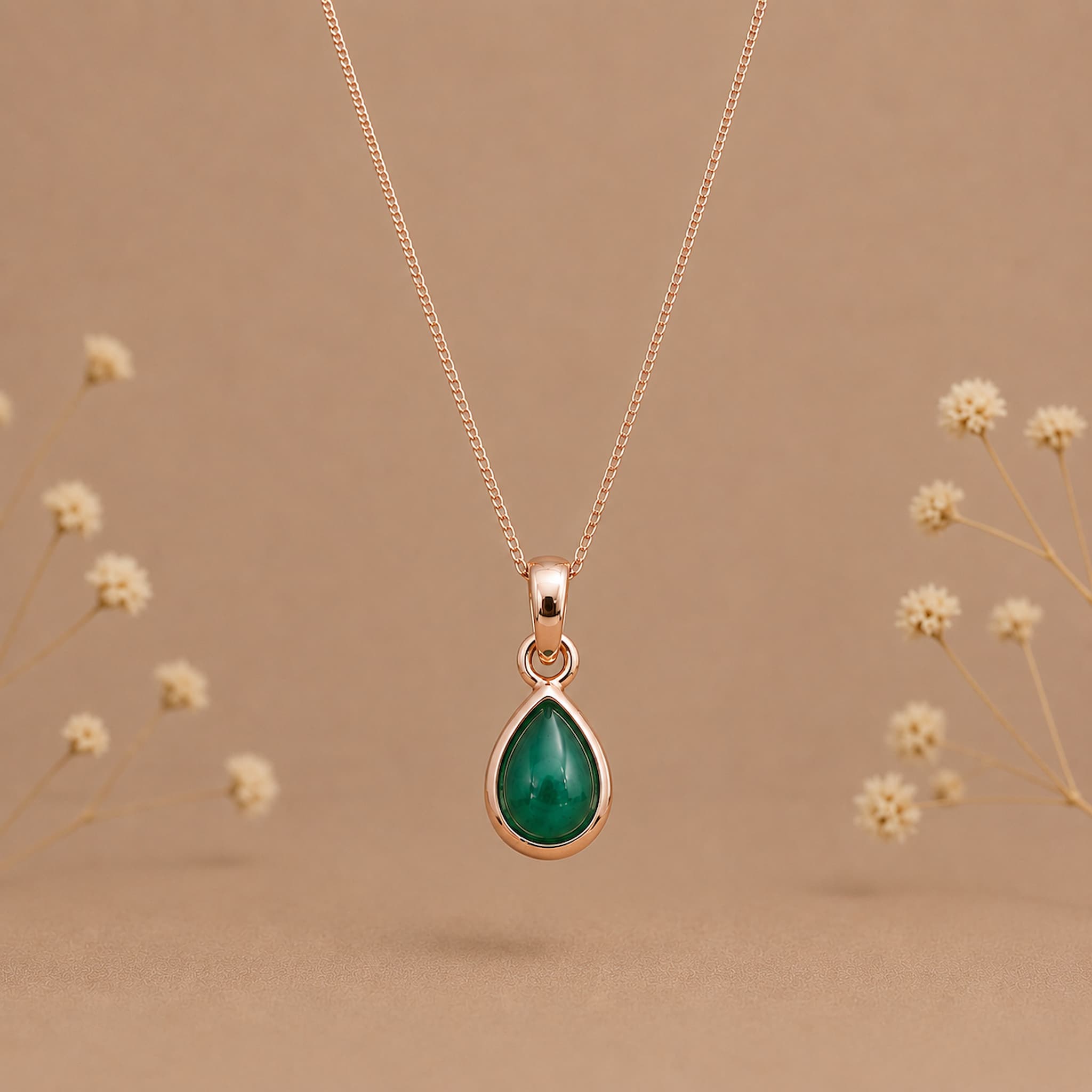 A rose gold may birthstone necklace with a teardrop pendant necklace with a natural green emerald gemstone, displayed against a warm beige background with small blurred dried flowers.