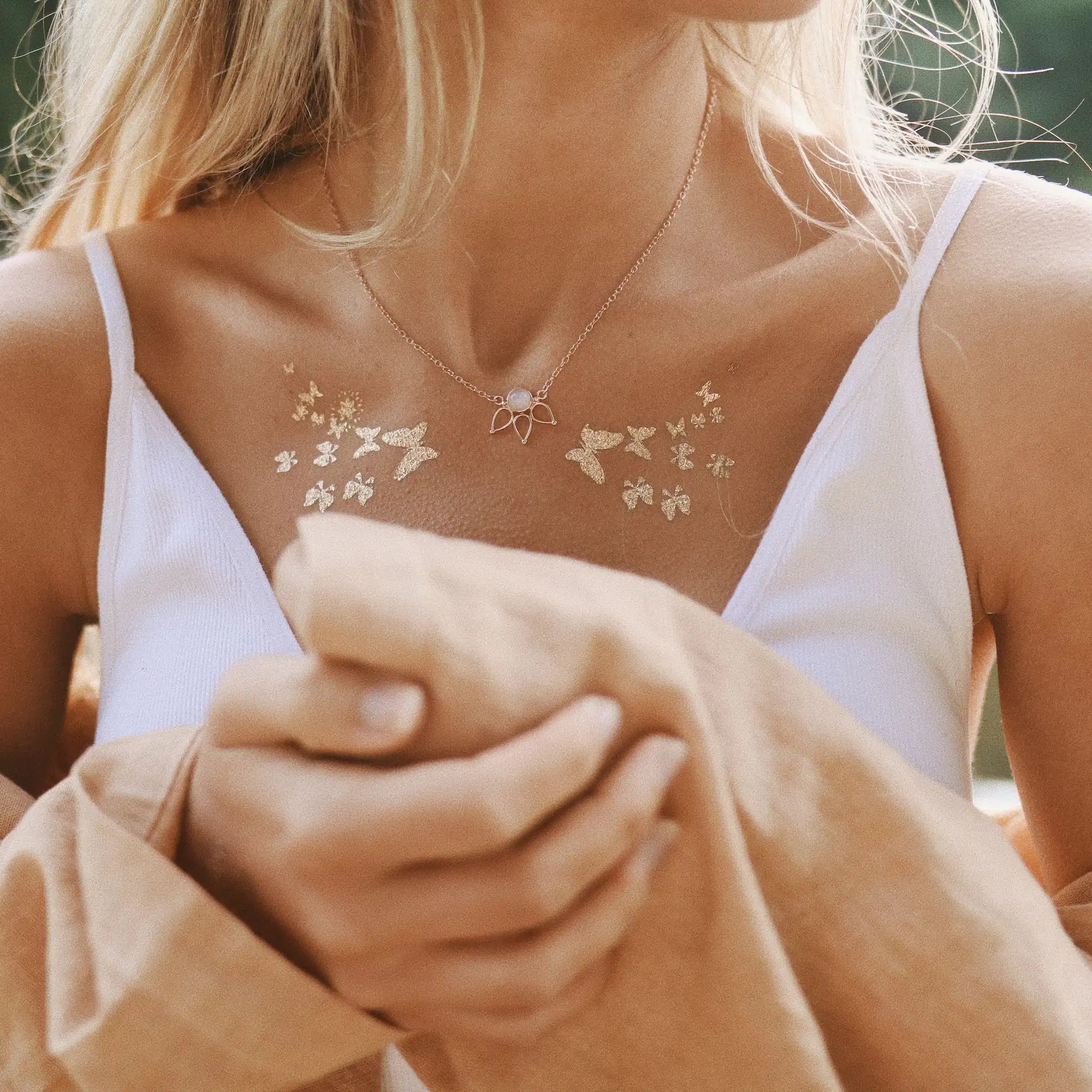 A close-up lifestyle shot of a woman wearing a delicate gold floral necklace with a small moonstone pendant, styled with a white camisole and a beige jacket.