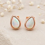 Pair of Rose Gold October Birthstone Earrings in pear-shaped cabochon stud style with natural opal gemstones on a light surface.