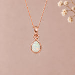 A rose gold october birthstone necklace featuring a teardrop pendant with a iridescent natural opal gemstone, suspended from a delicate rose gold chain against a soft beige background with blurred dried flowers on the right.
