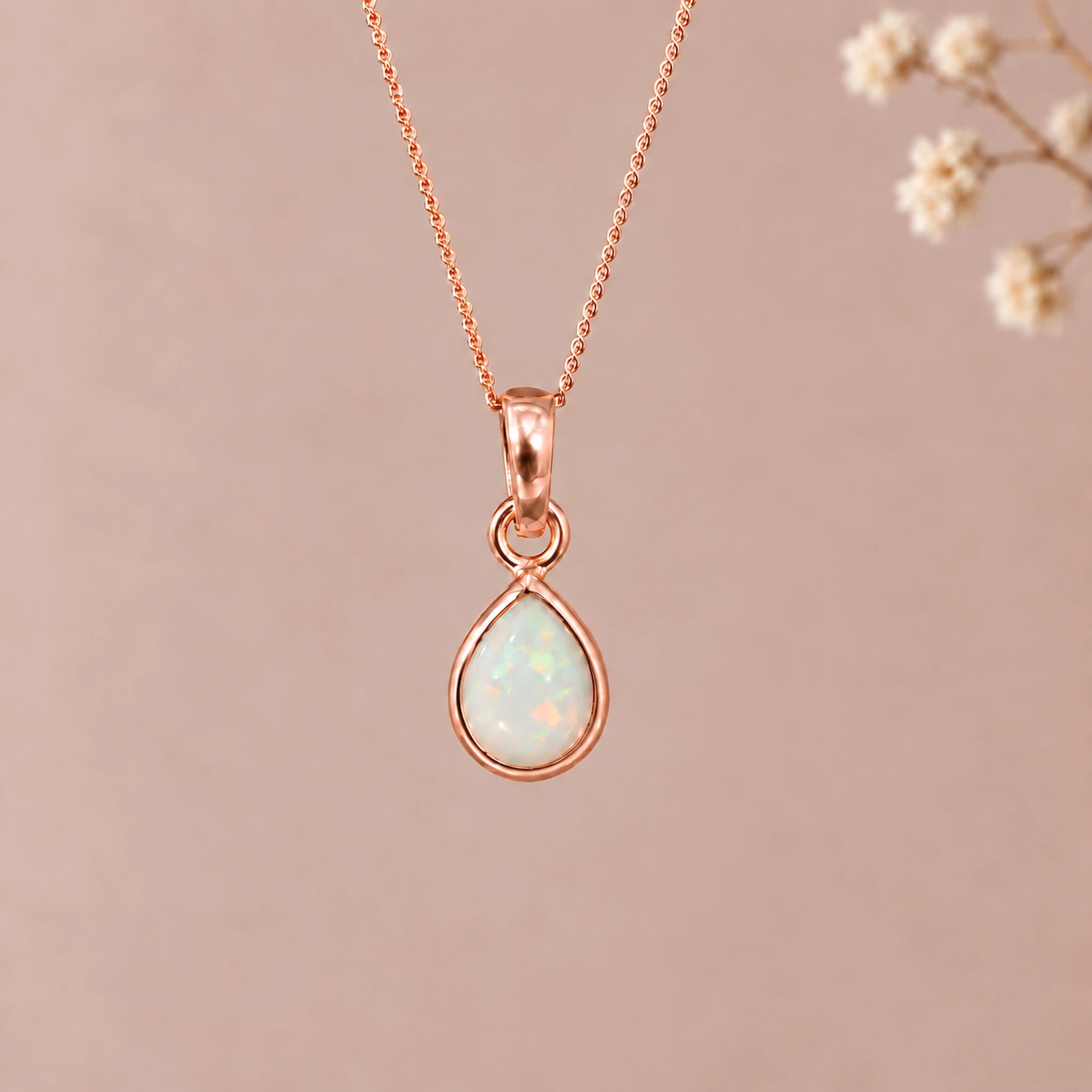 A rose gold october birthstone necklace featuring a teardrop pendant with a iridescent natural opal gemstone, suspended from a delicate rose gold chain against a soft beige background with blurred dried flowers on the right.