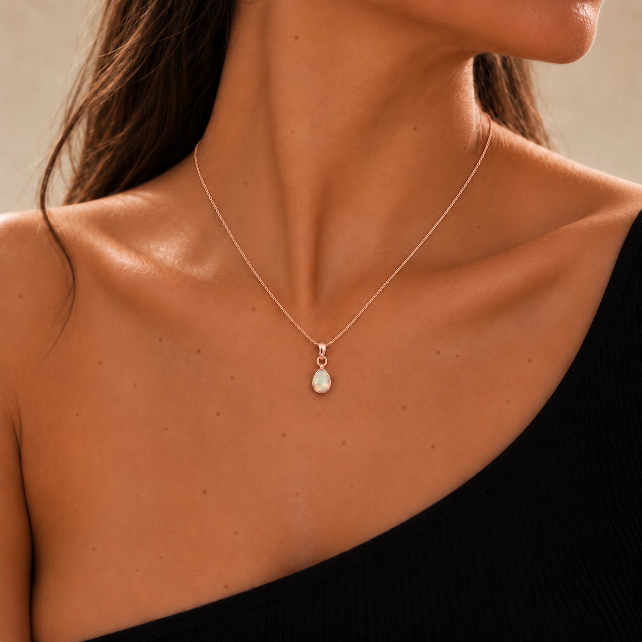 A close-up of a model's neck and upper chest wearing a delicate rose gold chain october birthstone necklace with a teardrop-shaped natural white opal pendant.