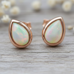 Teardrop-shaped opal earrings with rose gold setting on a wooden surface with blurred flowers in the background.