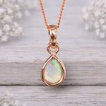Rose gold pendant with a teardrop-shaped opal stone on a wooden surface with blurred white flowers in the background.