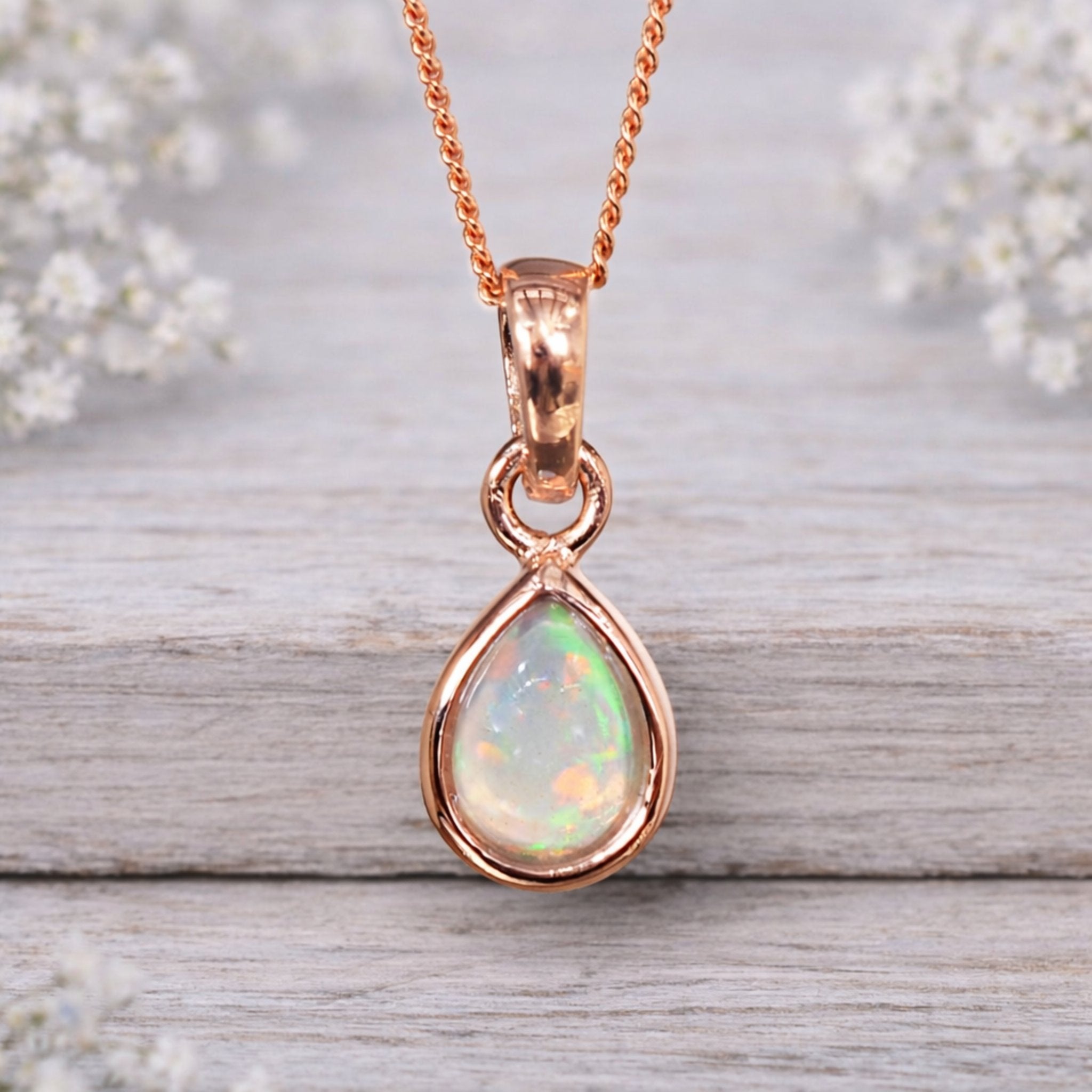 Rose gold pendant with a teardrop-shaped opal stone on a wooden surface with blurred white flowers in the background.