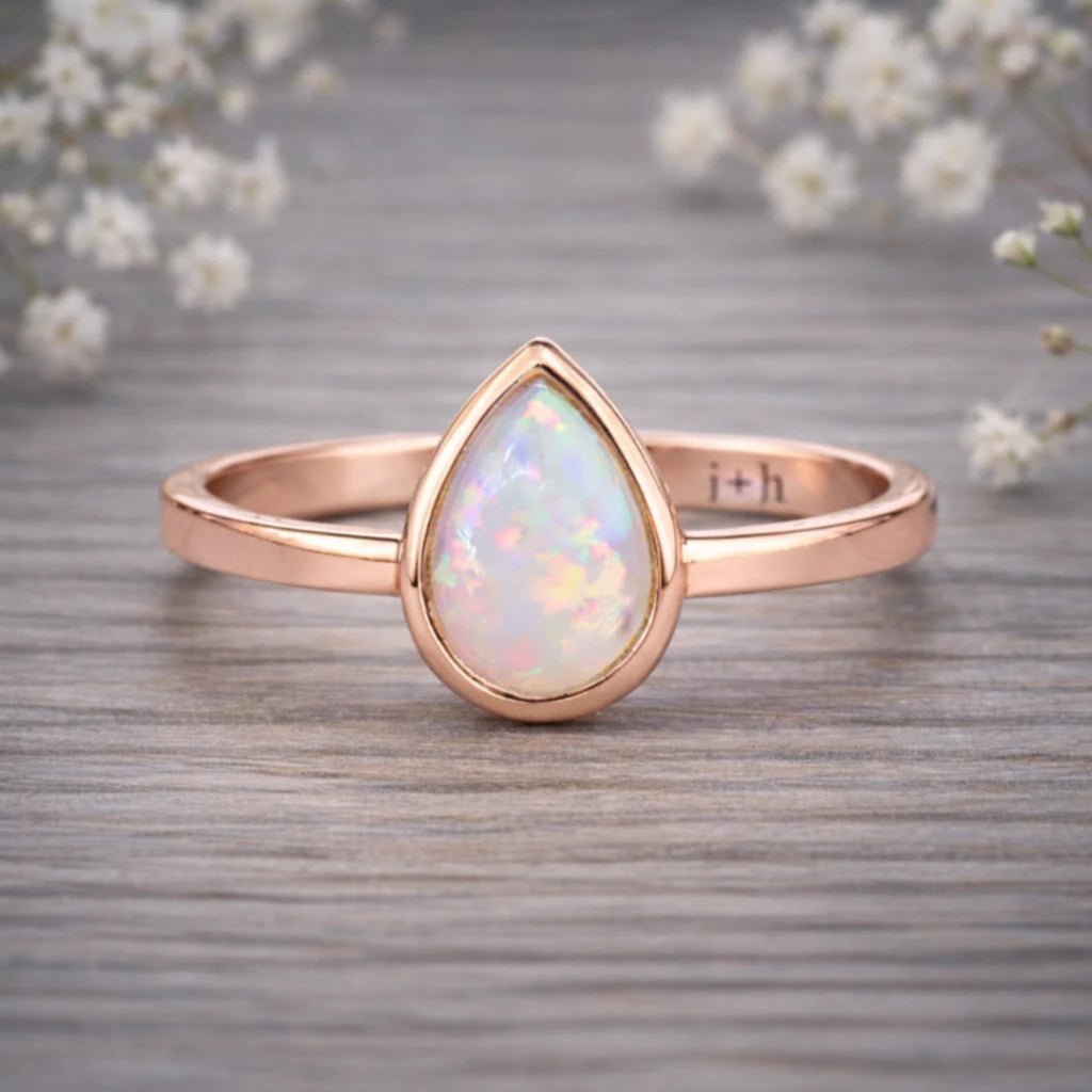 Rose gold ring with a teardrop-shaped opal stone on a wooden surface with white flowers in the background.