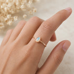 A delicate gold ring with a teardrop-shaped light blue opal gemstone, worn on a person's ring finger against a soft, warm neutral background with dried flowers.