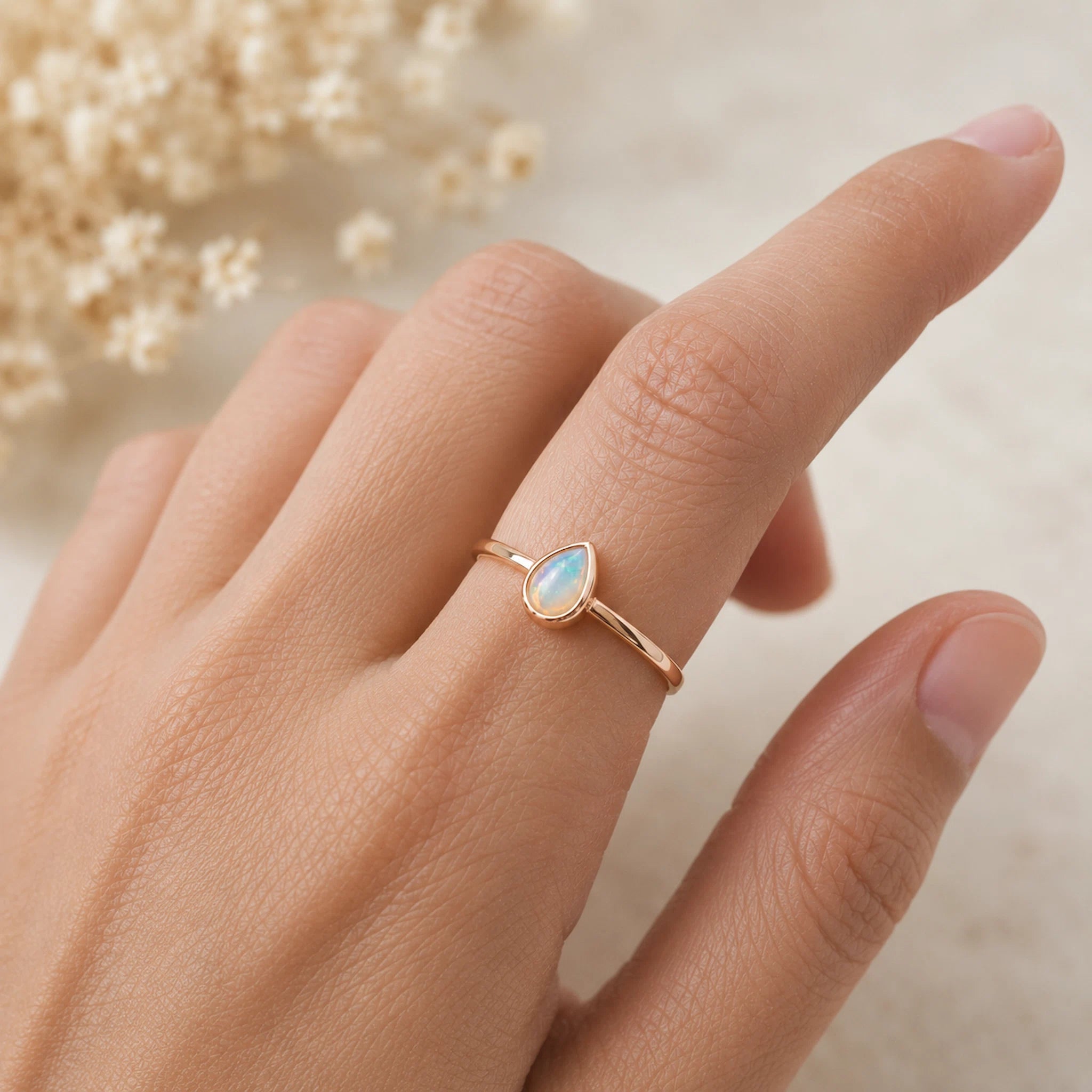 A delicate gold ring with a teardrop-shaped light blue opal gemstone, worn on a person's ring finger against a soft, warm neutral background with dried flowers.