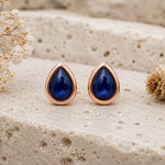 A pair of September Birthstone Natural Sapphire Earrings - teardrop-shaped stud earrings with deep blue stones and rose gold settings, displayed on a textured beige stone surface with dried flowers.