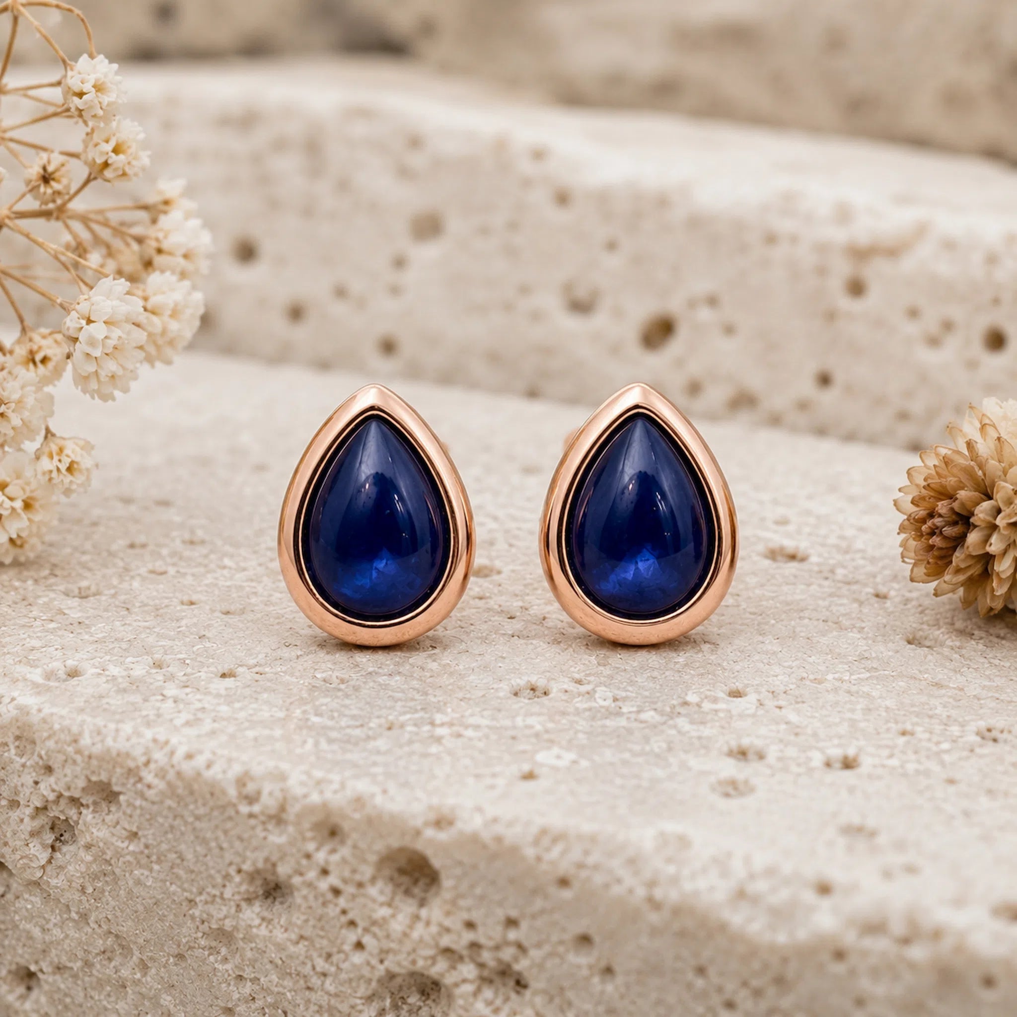 A pair of September Birthstone Natural Sapphire Earrings - teardrop-shaped stud earrings with deep blue stones and rose gold settings, displayed on a textured beige stone surface with dried flowers.