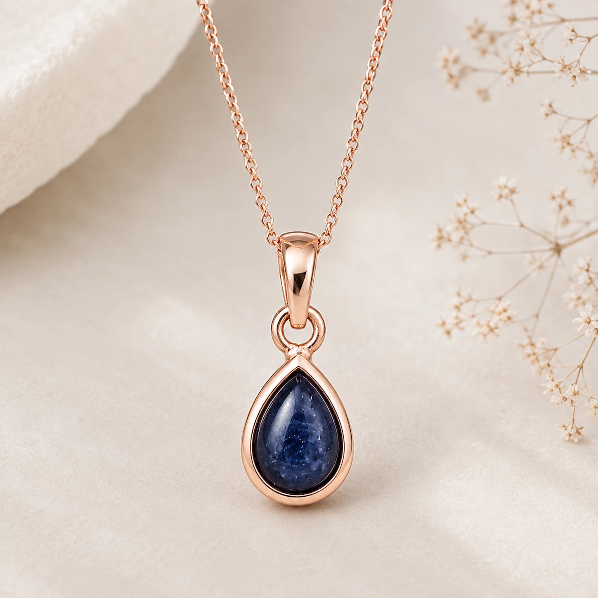 September Birthstone Natural Sapphire Necklace - A rose gold pendant necklace featuring a teardrop-shaped dark blue sapphire stone, displayed on a soft cream fabric background with delicate dried baby's breath flowers.
