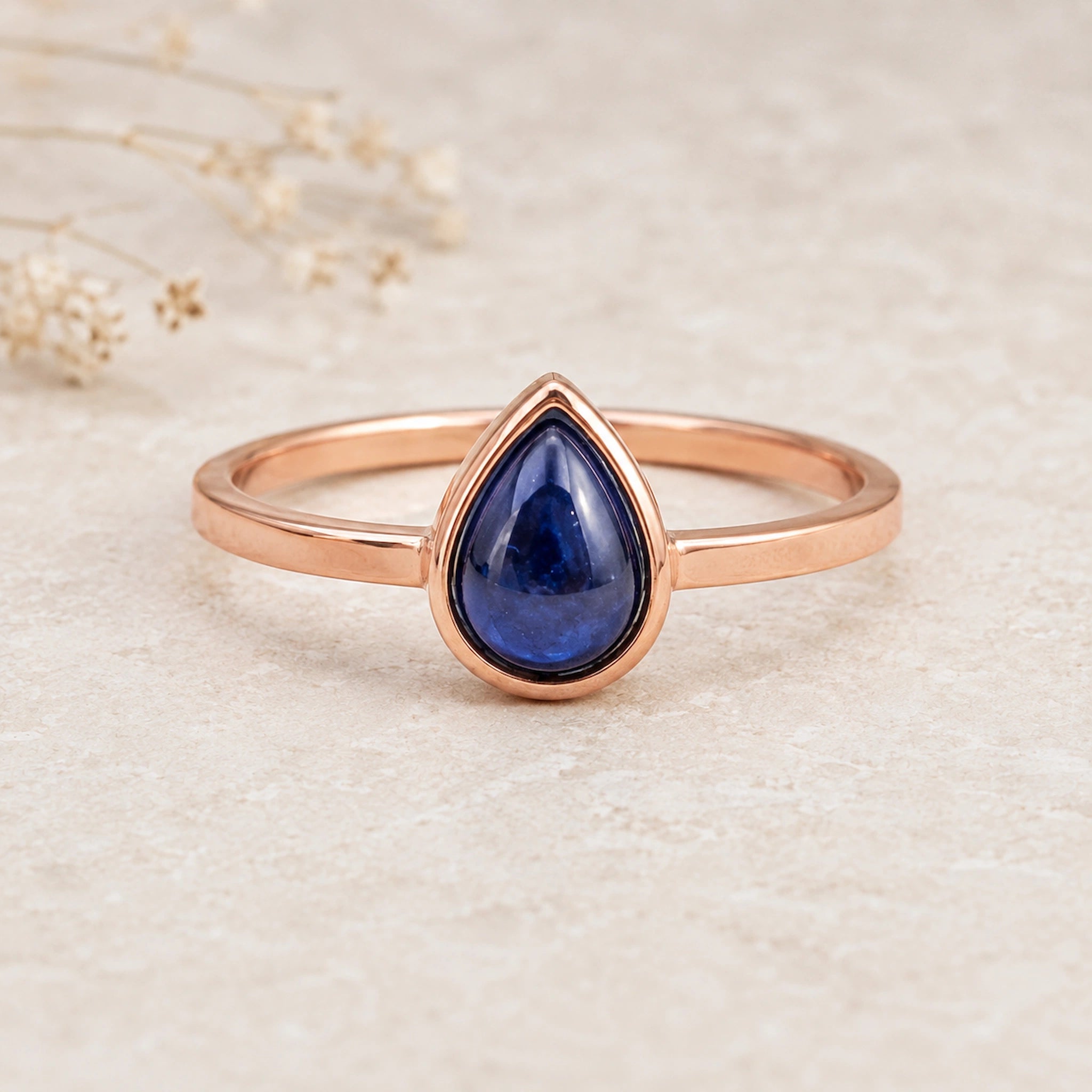 September birthstone Natural Sapphire Ring - A rose gold ring with a teardrop-shaped dark blue gemstone, displayed on a light beige textured surface with dried flowers in the background.