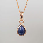 Teardrop-shaped blue sapphire gemstone pendant on a rose gold chain against a gray background