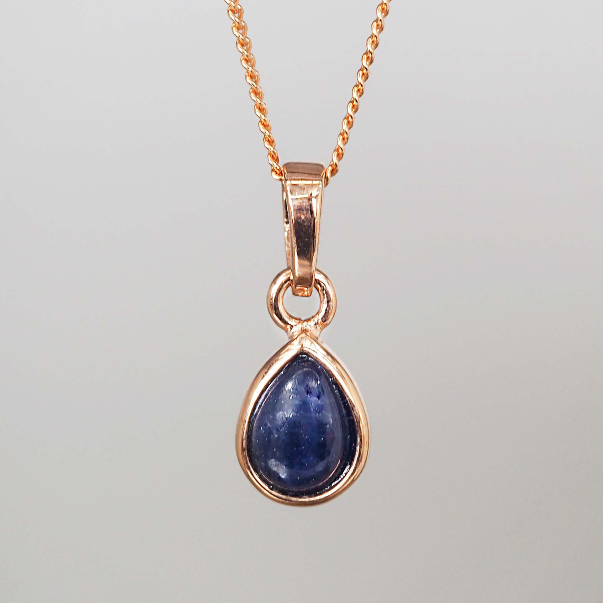 Teardrop-shaped blue sapphire gemstone pendant on a rose gold chain against a gray background