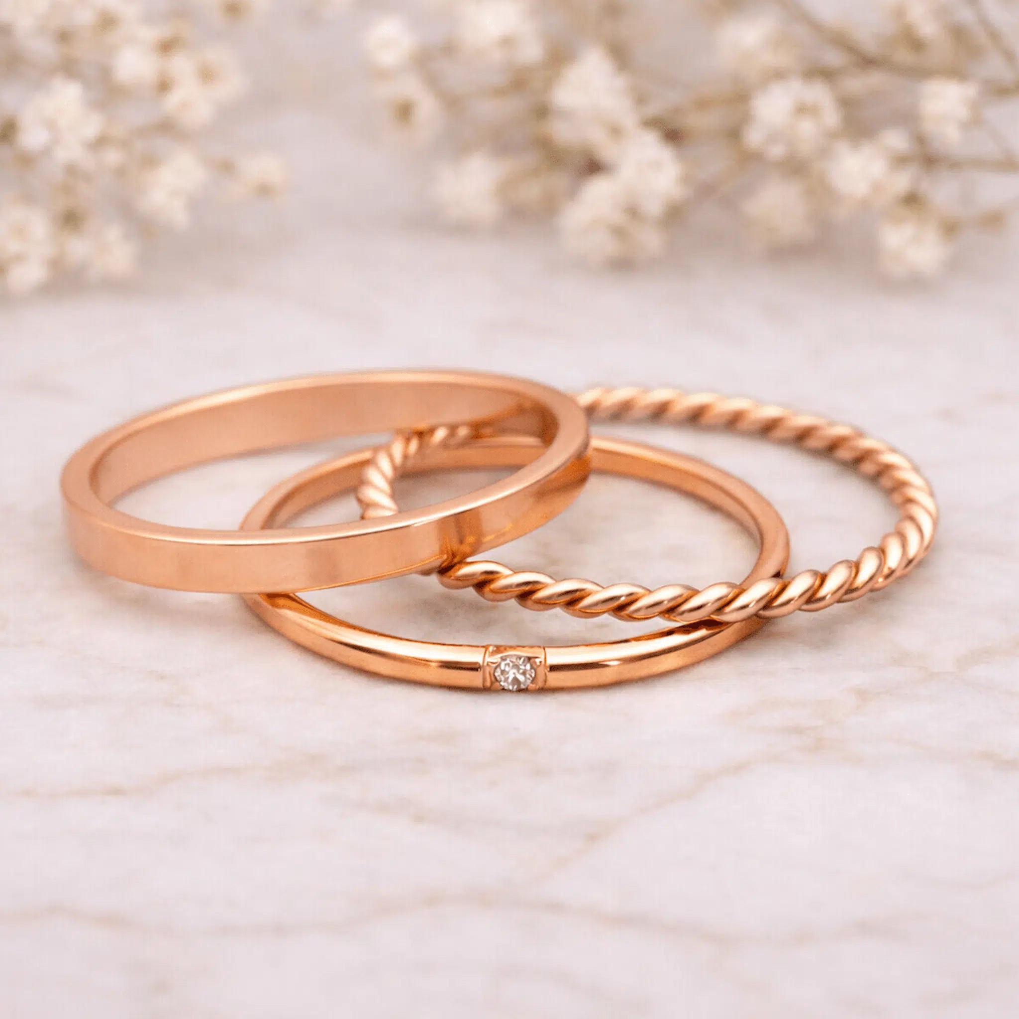 Three rose gold stacker rings on a marble surface with blurred flowers in the background