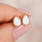 A pair of rose gold teardrop stud earrings with white opal inlays, held between fingers with a soft pink manicure against a warm beige background.