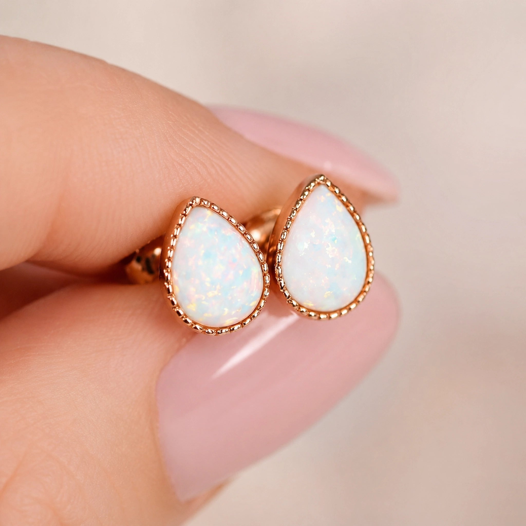 A pair of rose gold teardrop stud earrings with white opal inlays, held between fingers with a soft pink manicure against a warm beige background.