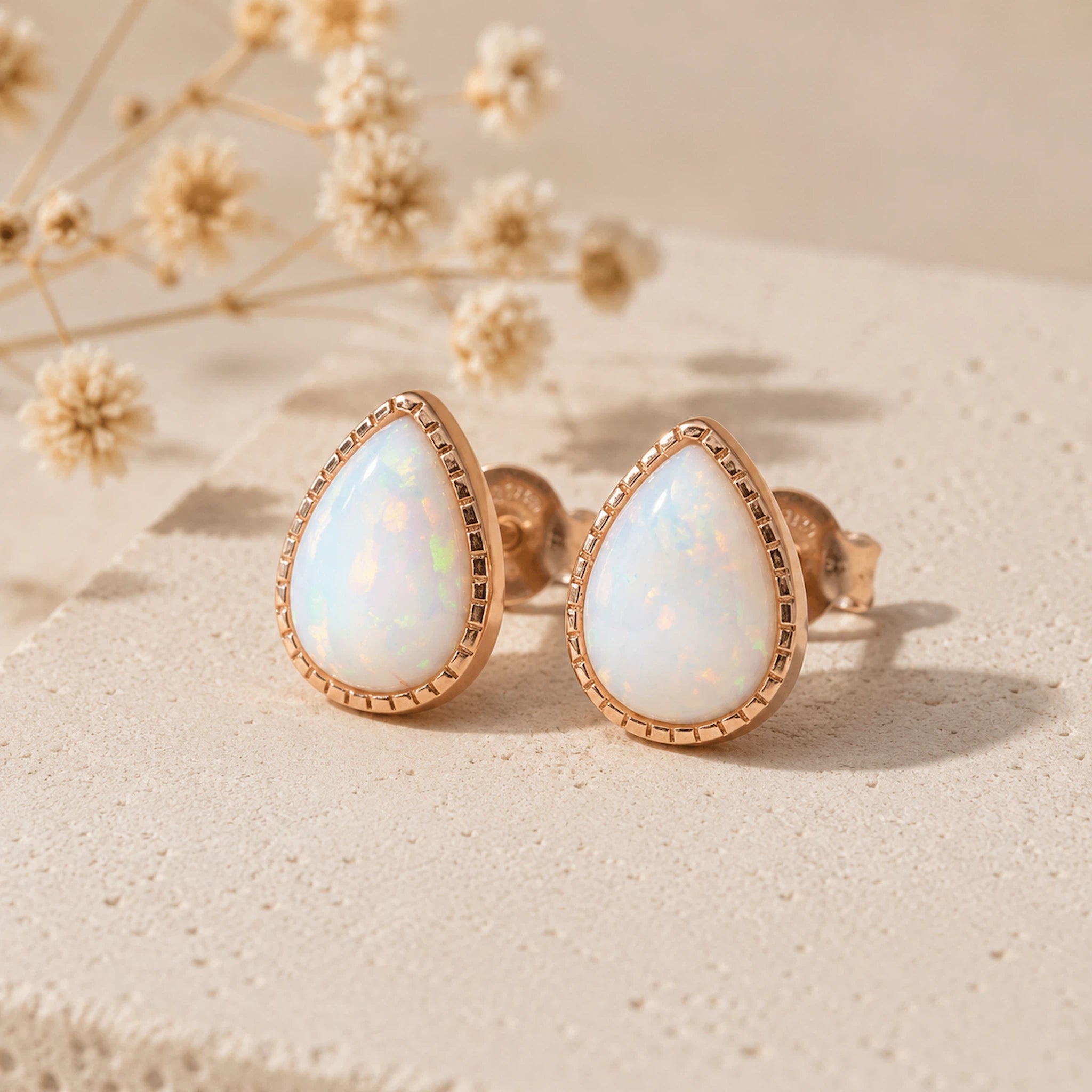 A pair of teardrop-shaped stud earrings with iridescent white opal centers and rose gold bezels, displayed on a textured beige surface with dried flowers in the background.
