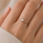 Womans hand wearing a minimalist thin Rose gold ring with a bezel set lab-create white opal stone against a neutral background