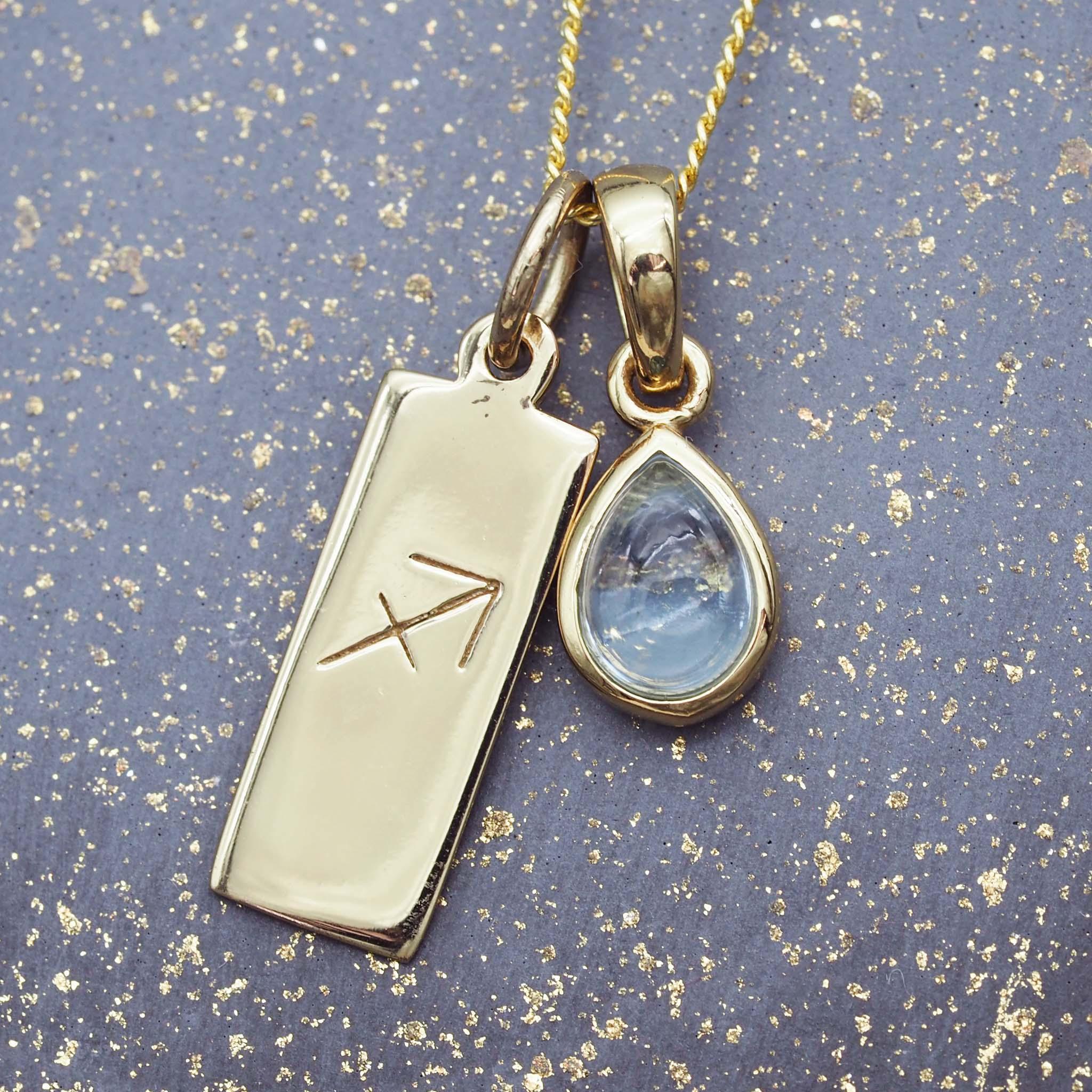 Gold necklace with a sagittarius zodiac pendant and a teardrop-shaped blue topaz gemstone on a gold and blue speckled background.
