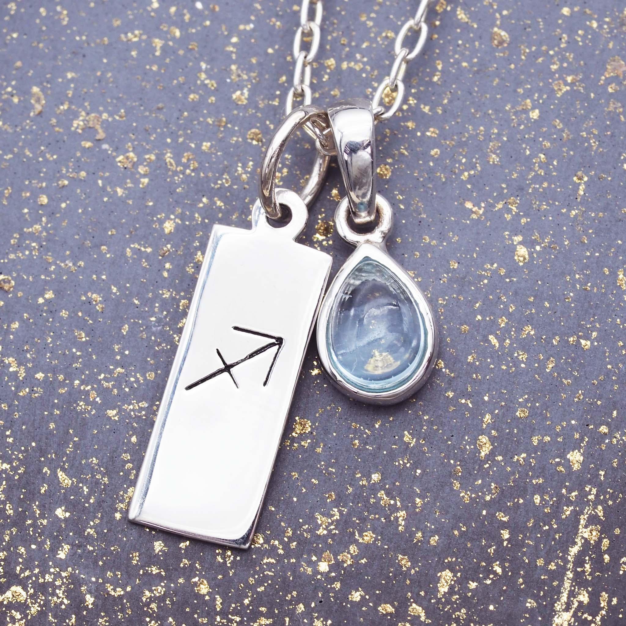 Silver necklace with a sagittarius zodiac pendant and teardrop-shaped topaz gemstone pendant on a textured gray background.