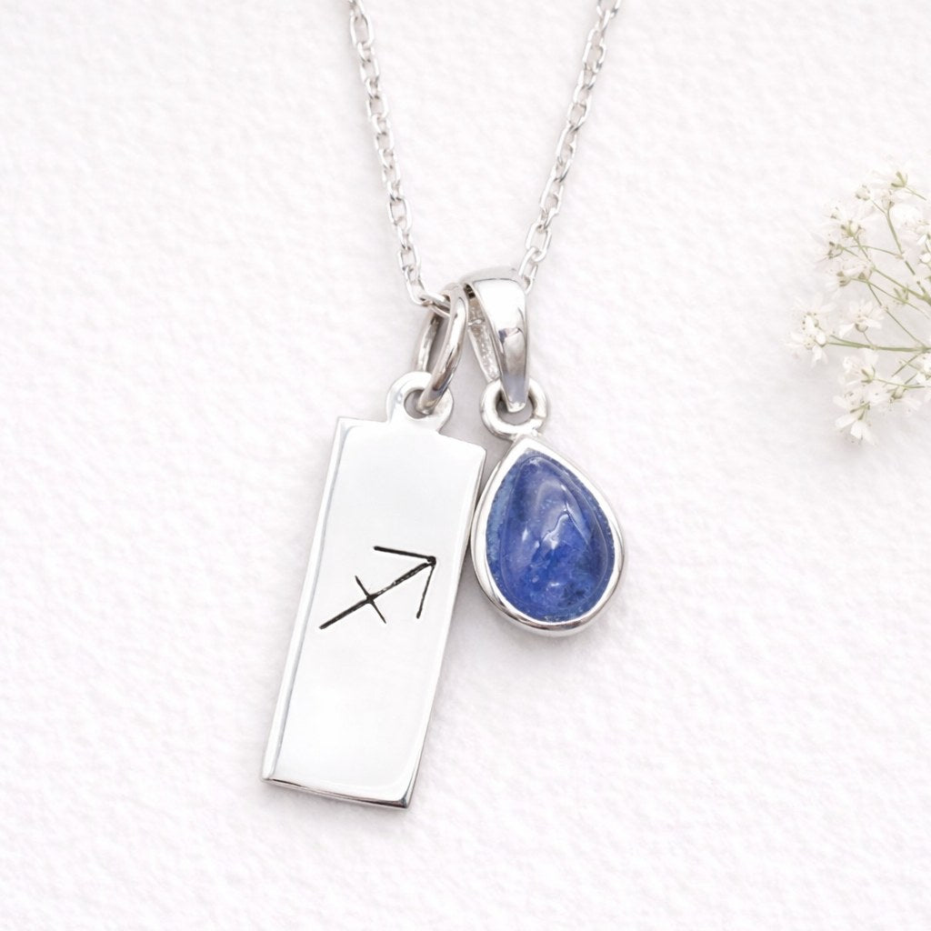 Silver necklace with a rectangular pendant with the sagittarius zodiac symbol and teardrop-shaped blue natural tanzanite gemstone pendant on a textured white background.