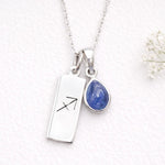 Silver necklace with a rectangular pendant with the sagittarius zodiac symbol and teardrop-shaped blue natural tanzanite gemstone pendant on a textured white background.